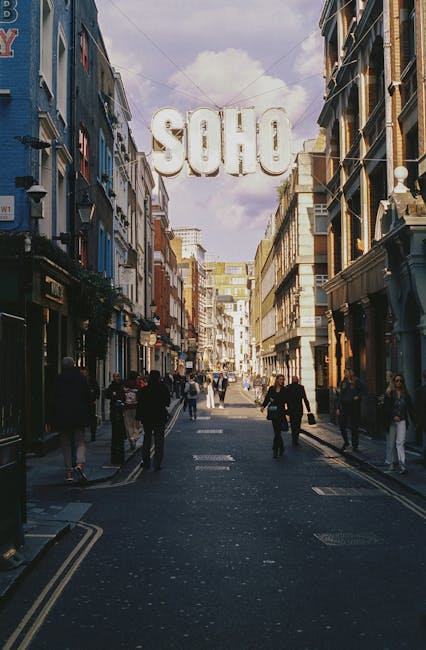 A narrow city street in Soho W1D, with brick and modern building facades on either side, decorated with hanging orange lanterns. The pavement is made of dark bricks, with red bollards lining the edge