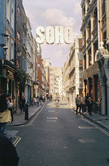 A busy street scene on Shaftesbury Avenue in Soho, featuring narrow pavements lined with multi-story retail and residential buildings with traditional architectural details, including large windows an