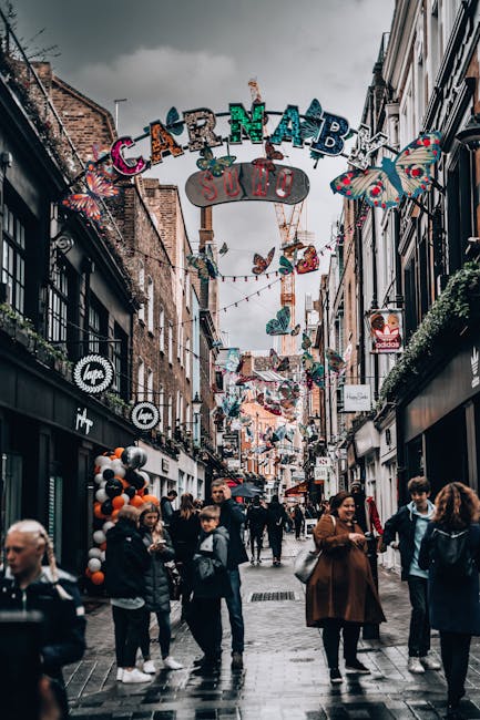 A busy street scene on Carnaby Street in Soho during daylight, with numerous pedestrians walking along the pavement, some carrying shopping bags. Overhead, colorful decorations and banners, including a large sign reading 'Carnaby,' hang across the narrow street, which is lined with retail stores featuring displays and signage. Shops on either side have large windows showcasing merchandise, with some storefronts decorated with balloons and festive materials. The environment appears lively and vibrant, with a mix of people browsing stores and engaging in conversation. The cloudy sky above suggests overcast weather, while the street is illuminated by natural light. This setting illustrates the typical busy and decorative atmosphere of a popular shopping and leisure area in London, often associated with recommendations for home relocation or furniture transport through services like Movers Soho, particularly when planning a move or packing during a busy day of errands in Soho.
