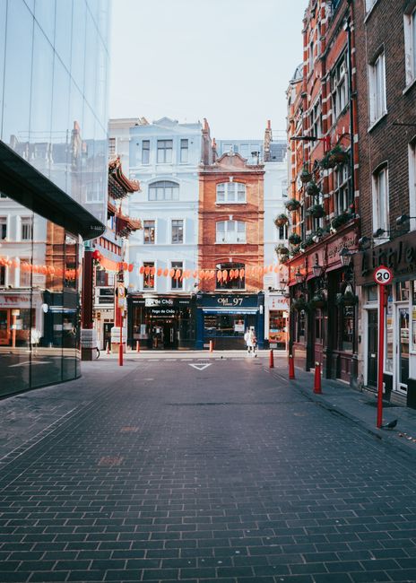 A paved pedestrian street in Soho, London, with a mix of historic and modern buildings on either side. On the left, a glass-fronted building reflects the surroundings, while on the right, brick facades feature windows with flower boxes and signage for local businesses. String lights are hung across the street, adding a festive atmosphere. In the background, several multi-storey buildings with classic architectural details are visible, with some windows open and chimney stacks on rooftops. Street bollards and red plastic barriers mark the boundary of the pedestrian zone. This scene captures the typical urban environment where house removals and furniture transport may occur, with no people visible but an indication of an area used for moving logistics, fitting the context of a Soho removals guide from Carnaby Street to Tottenham Court Road, as provided by Movers Soho.