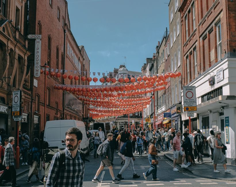 A busy street scene on Carnaby Street in Soho during daylight, with numerous pedestrians walking along the pavement, some carrying shopping bags. Overhead, colorful decorations and banners, including a large sign reading 'Carnaby,' hang across the narrow street, which is lined with retail stores featuring displays and signage. Shops on either side have large windows showcasing merchandise, with some storefronts decorated with balloons and festive materials. The environment appears lively and vibrant, with a mix of people browsing stores and engaging in conversation. The cloudy sky above suggests overcast weather, while the street is illuminated by natural light. This setting illustrates the typical busy and decorative atmosphere of a popular shopping and leisure area in London, often associated with recommendations for home relocation or furniture transport through services like Movers Soho, particularly when planning a move or packing during a busy day of errands in Soho.